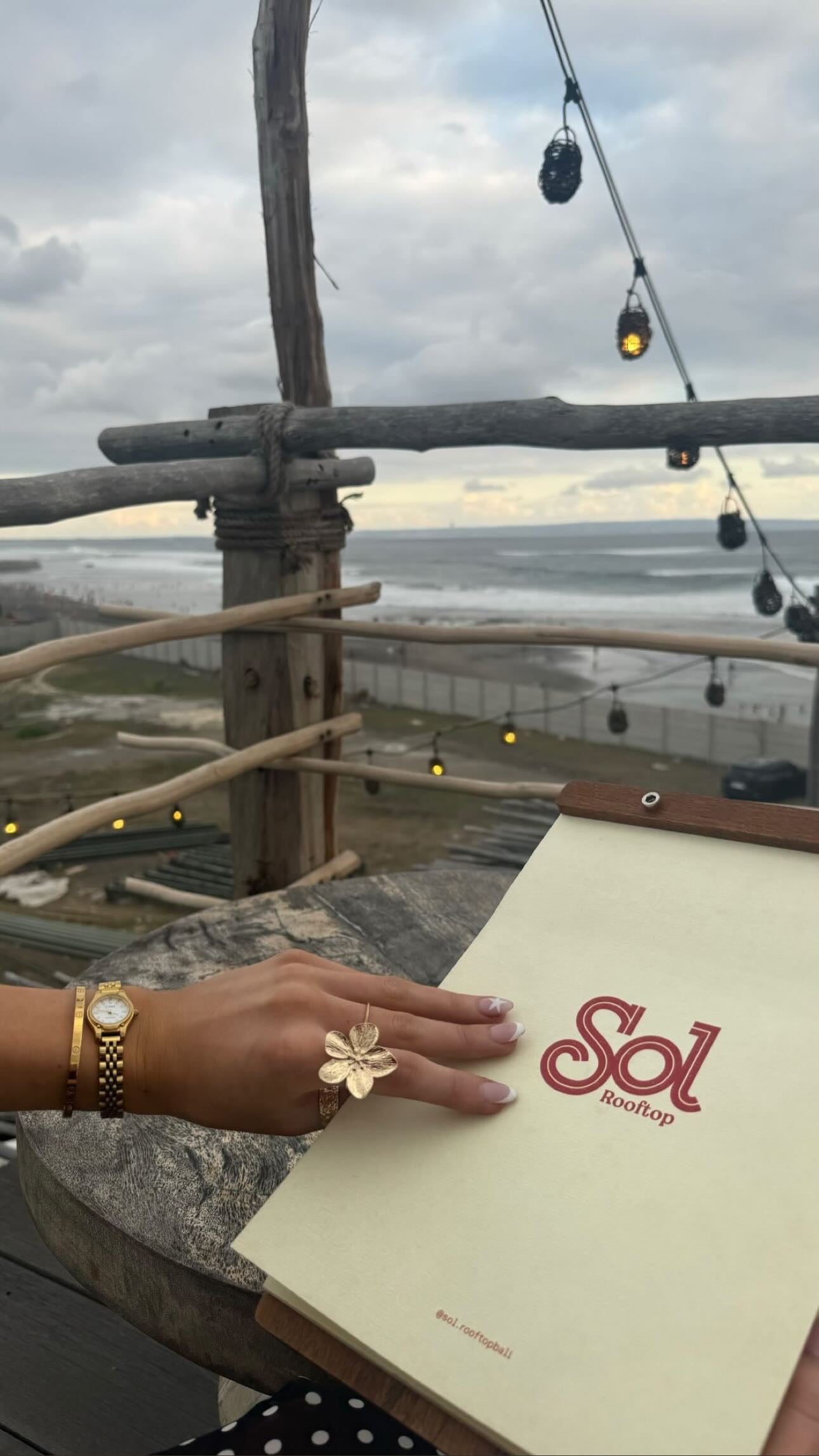 Person's hand wearing the Fleur Ring, a stunning flower statement ring by ÈRE, while holding a menu at a rooftop venue, showcasing its elegance in lifestyle settings.